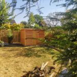 new small shed in the left corner of a lawned garden surrounded by trees, Single door that is closed with pent roof.
