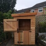 narrow new timber small shed with pent roof with stable doors on the narrow end, the top one is open with a flower bed on the right
