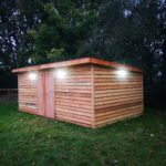 a new large timber building built in a field with pent roof with three lights on under lighting up the sides in the daylight